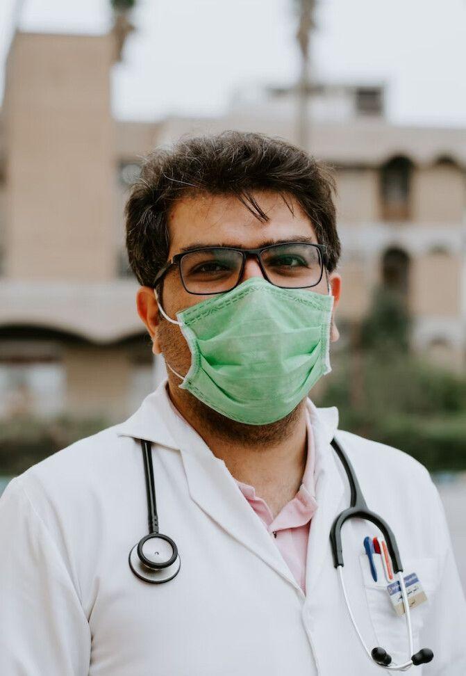 A doctor wearing a green face mask and glasses stands outdoors. He is dressed in a white coat with a stethoscope around his neck and pens in his pocket, suggesting he is ready for a medical consultation. The background shows a building, indicating he might be near a hospital or clinic.