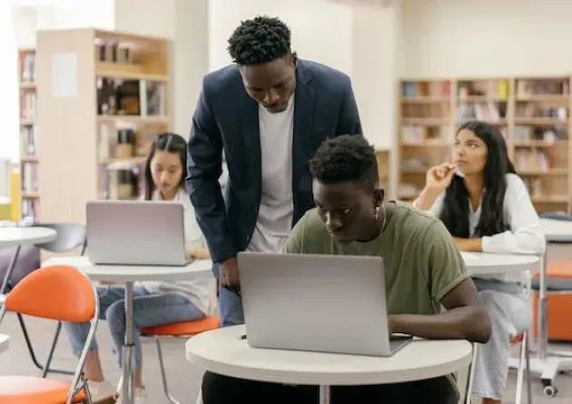 An ESL teacher assists a student working on a laptop in a classroom. Other students are also engaged with their laptops, creating a focused learning environment.