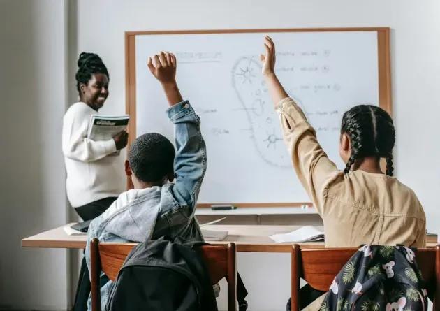 A high school teacher smiles as two students raise their hands to answer a question during a biology lesson. The whiteboard behind them shows a diagram and notes, creating an interactive classroom environment.